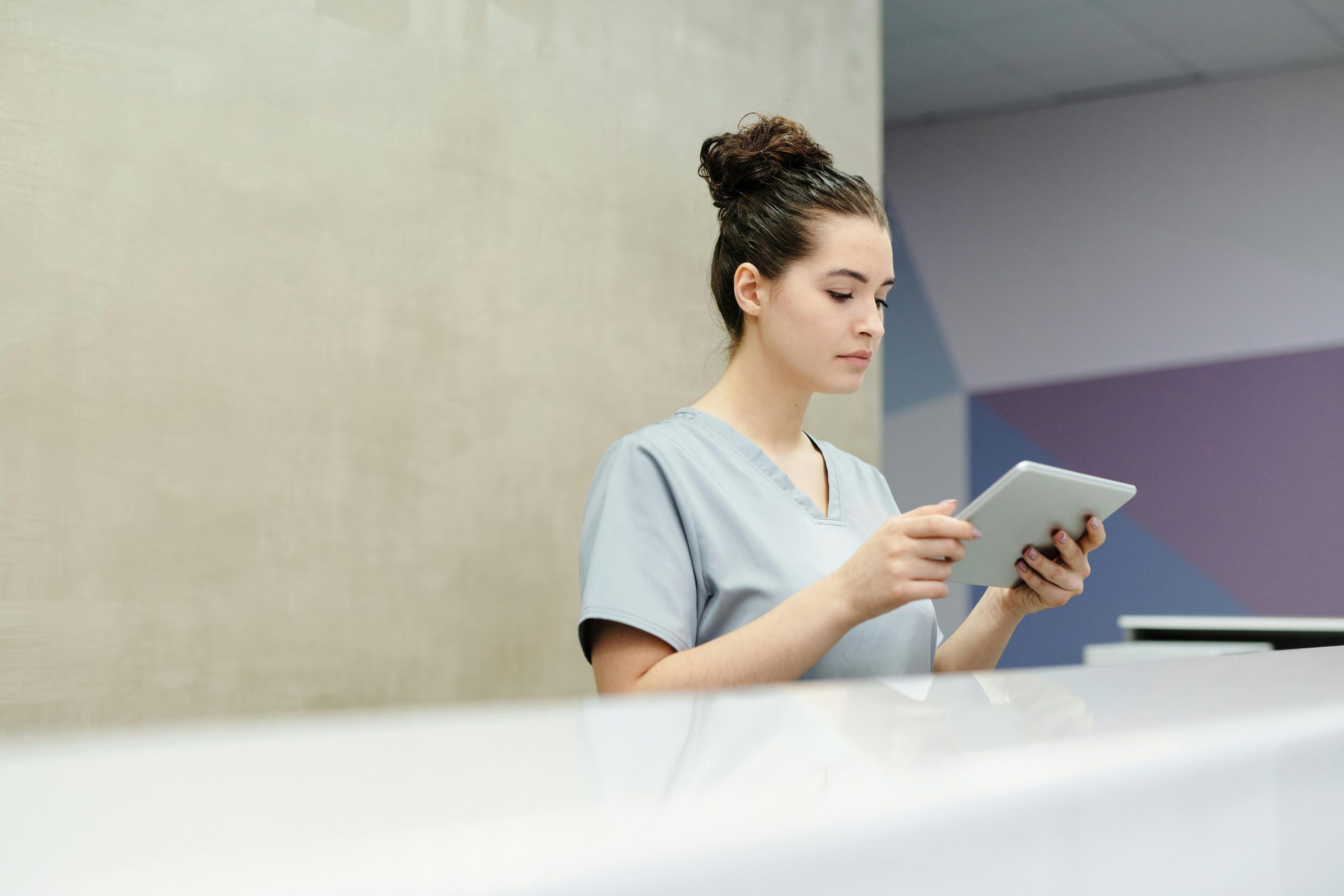 A receptionist in scrubs using a tablet at an office reception desk.